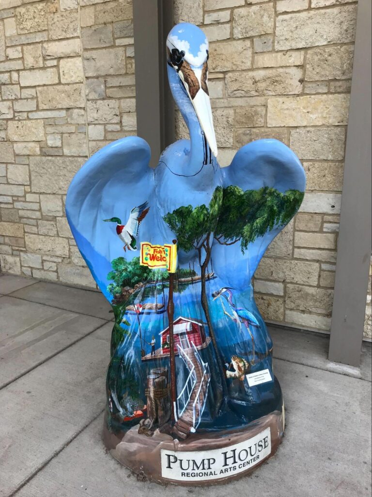 A 6’ tall heron sculpture with a landscape painted on the sculpture including water, a brown dock, a red boat house, green trees in the background, and a Mallard duck flying through the blue sky.