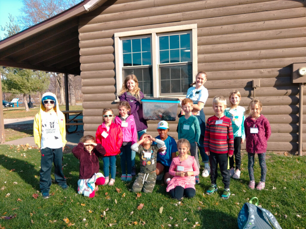 A group of 11 homeschool students are posing outdoors. Behind them, two Environmental Educators are holding a plastic tote box with a rotten pumpkin inside. 
