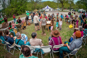 Community members sit in a large circle of chairs placed outside The Nature Place and play drums with Drums For Peace at the 2016 Earth Fair.