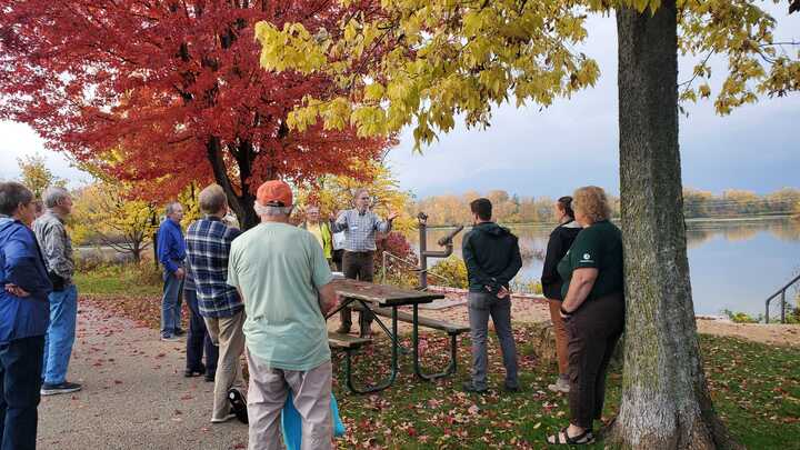 Adults stand outside listening to a speaker talk. The tree on the upper left side has bright red and yellow autumn leaves and the ground is green grass covered in leaves.