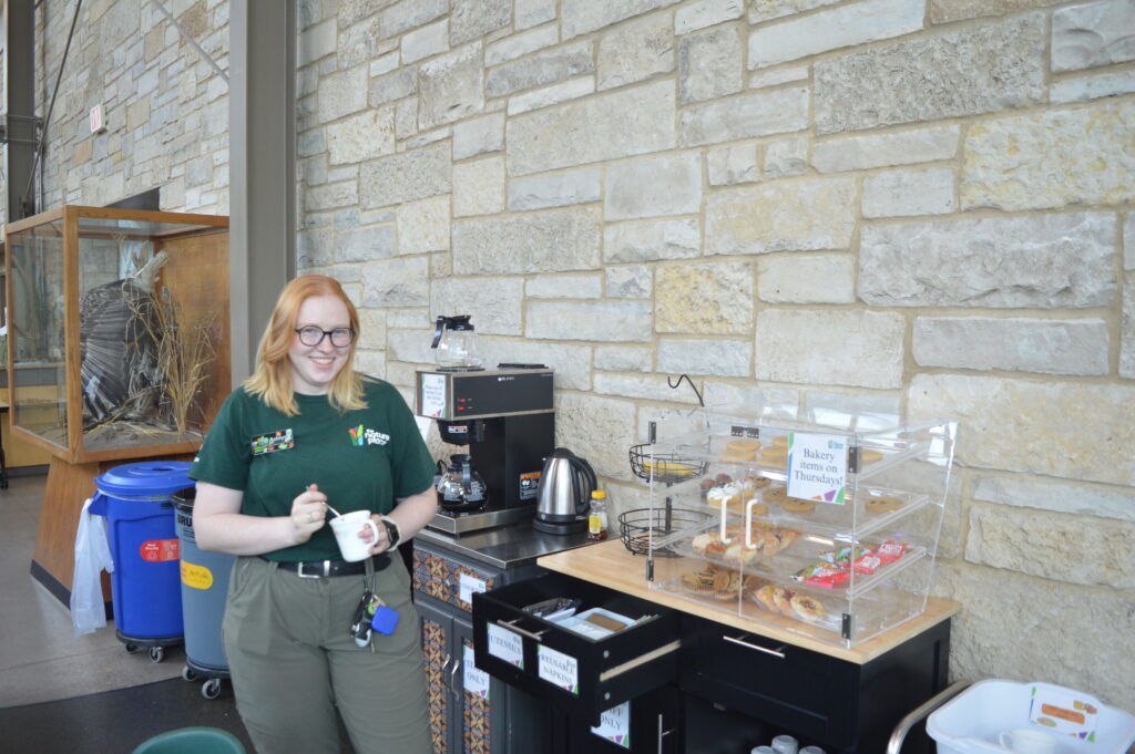 Young woman with red orange hair and a green shirt holding a coffee mug. There is a coffee bar behind her, as well as pastries. 