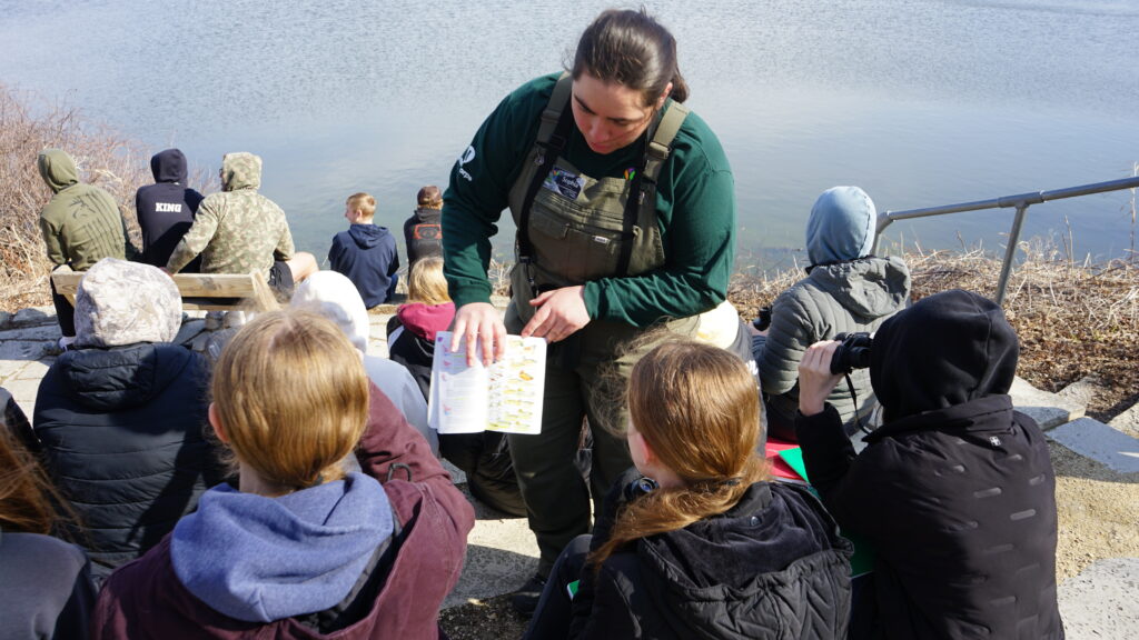A woman pointing to a book that kids are looking at in front of a body of water.