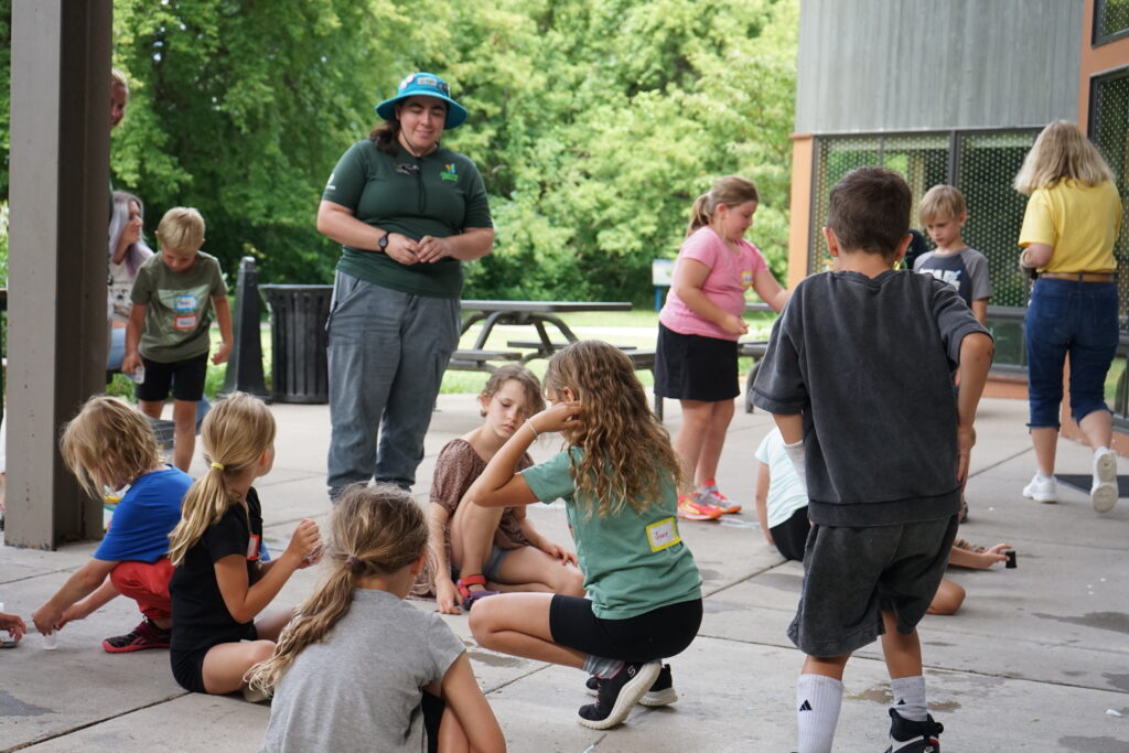 Group of Summer Day Campers at The Nature Place gather around AmeriCorps Member to complete a science experiment.